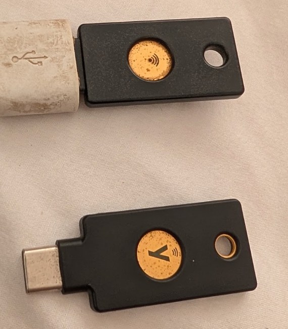 Two worn YubiKeys on a white sheet. On top, the USB-A YubiKey NEO, plugged into a white USB-A to USB-C adapter. Below it, the USB-C YubiKey 5 NFC. Both have the gold touch contact scuffed from years of use.